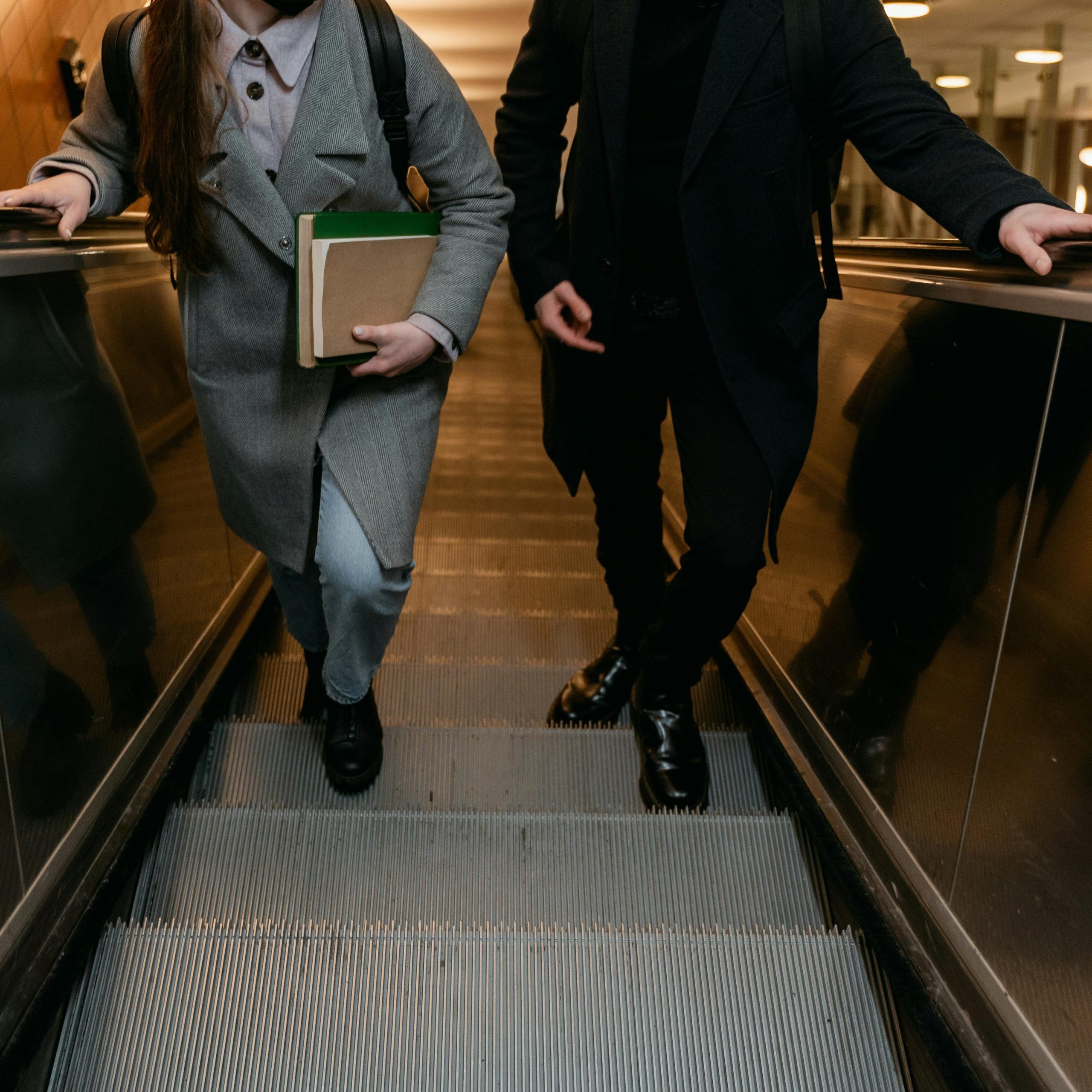 Two adults on an escalator indoors, holding books and bags.