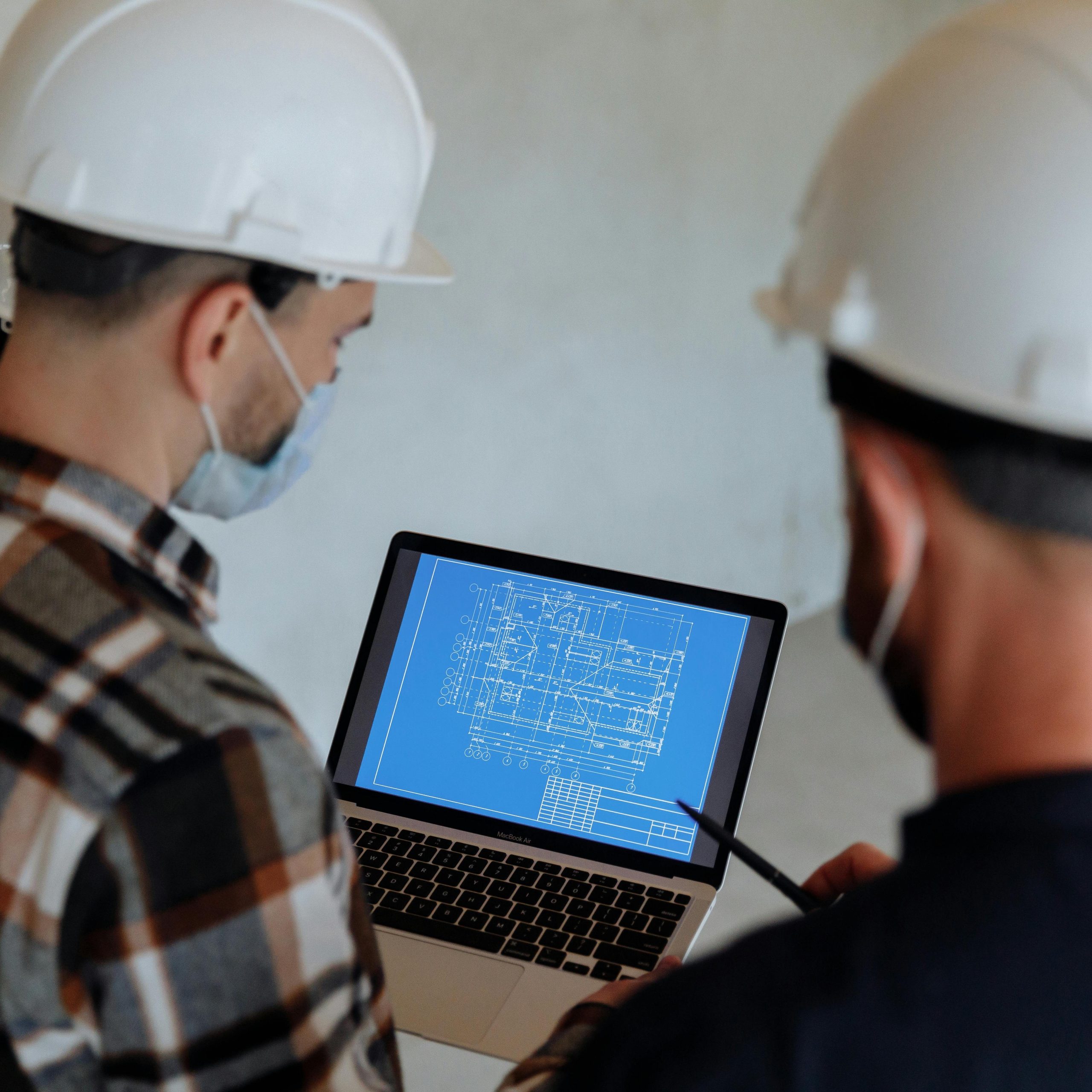 Engineers wearing hard hats and masks review a construction blueprint on a laptop indoors.