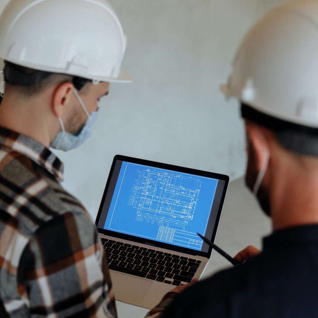 pexels-photo-8482865-8482865 Engineers wearing hard hats and masks review a construction blueprint on a laptop indoors.