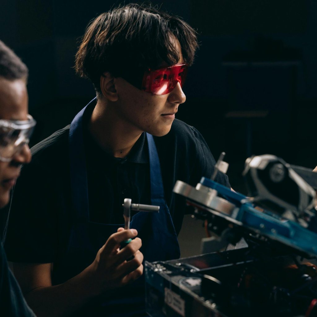 Two engineers wearing safety glasses operate machinery indoors, focused on mechanical tasks.