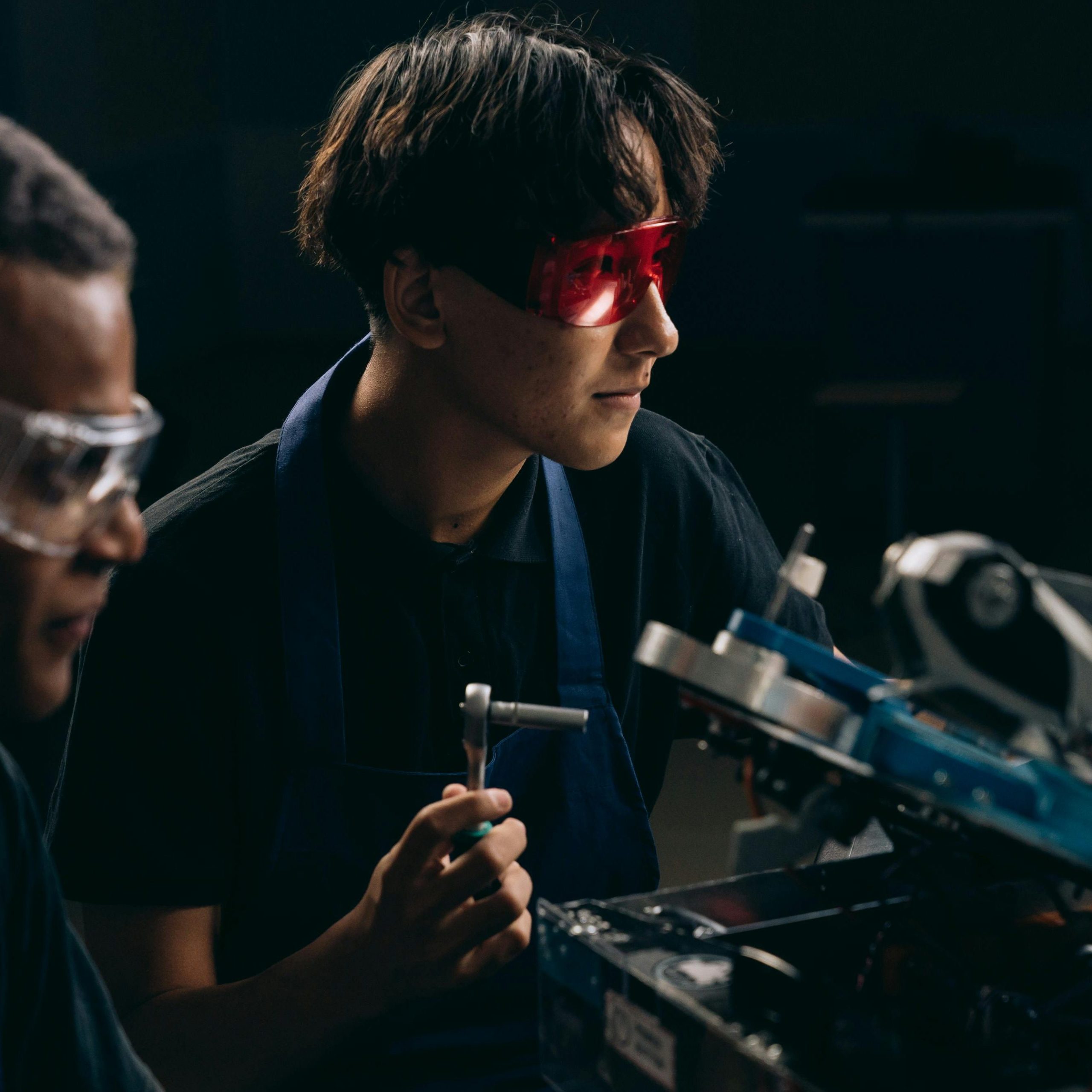 Two engineers wearing safety glasses operate machinery indoors, focused on mechanical tasks.